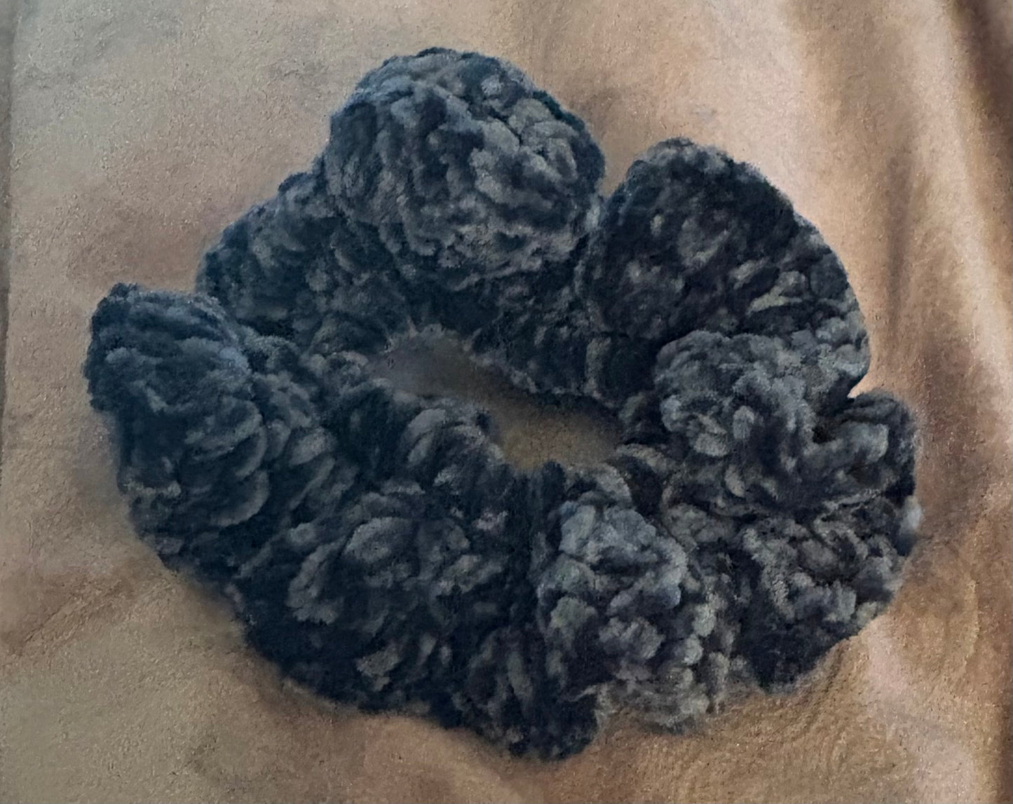 A dark gray, heavily textured and puffy scrunchie made of a plush, fuzzy fabric, resting on a beige fabric background.