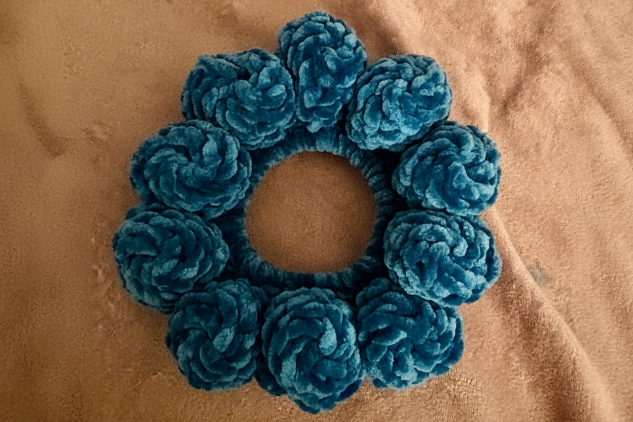 A blue textured scrunchie made of plush, rosette-like fabric loops, photographed from above on a tan textured surface.