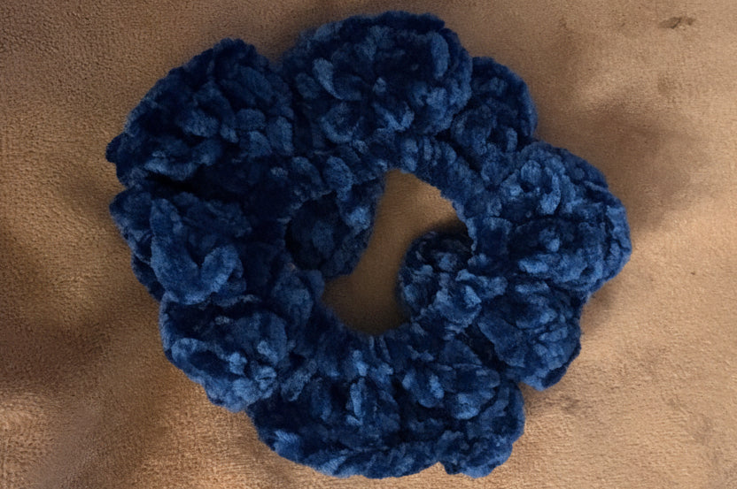 A single dark blue textured scrunchie made of chunky, plush terry cloth, placed on a brown suede-like surface.